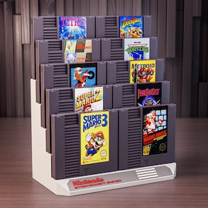 Könnte beinhalten: Ein weißer, abgestufter Präsentationsständer mit zehn grauen Nintendo Entertainment System-Spielkassetten. Die Etiketten zeigen Titel wie Tetris, DuckTales, Super Mario Bros. 2 und 3, Teenage Mutant Ninja Turtles, Metroid und Final Fantasy.
