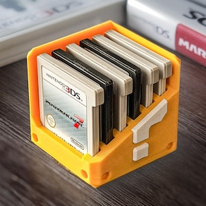 Puede incluir: Un soporte amarillo impreso en 3D para cartuchos de juego con un diseño de signo de interrogación. Contiene varios cartuchos de juego de Nintendo 3DS. Los cartuchos son negros, grises y blancos. El soporte está sobre una superficie de madera oscura.