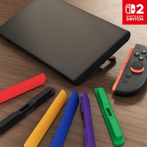 Könnte beinhalten: Eine schwarze Nintendo Switch-Konsole mit einem schwarz-orangenen Controller liegt auf einer Holzoberfläche. Bunte Controller-Abdeckungen in Rot, Schwarz, Gelb, Blau, Lila und Grün sind im Vordergrund angeordnet. Das Nintendo Switch-Logo befindet sich oben rechts.