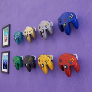 Puede incluir: Una colección de coloridos mandos de Nintendo 64 montados en una pared morada. Se muestran mandos en azul, verde, dorado, gris, negro, amarillo y rojo. También se ven dos cuadros enmarcados.