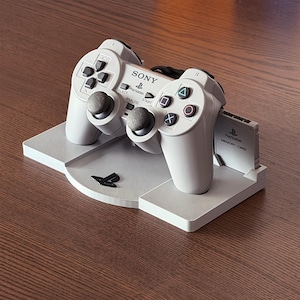 Op de afbeelding: Een lichtgrijze PlayStation-controller met zwarte knoppen en een geheugenkaart, rustend op een bijpassende standaard. De controller heeft de Sony- en PlayStation-logo's. De standaard heeft het PlayStation-logo.