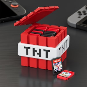 Puede incluir: Una caja de almacenamiento roja con temática TNT para cartuchos de juegos de Nintendo Switch, con la palabra "TNT" en blanco. La caja está abierta, revelando cartuchos de juego en su interior. Dos cartuchos de juego Switch están delante de la caja.