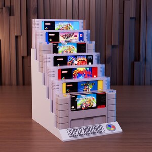May include: A white tiered display stand holding seven Super Nintendo Entertainment System game cartridges. The base features the text SUPER NINTENDO ENTERTAINMENT SYSTEM and a multi-coloured button logo. The cartridges are arranged in a stepped formation.