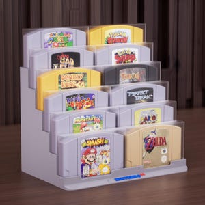 Könnte beinhalten: Ein abgestufter, hellgrauer Kunststoff-Präsentationsständer mit zehn Nintendo 64-Videospielkassetten. Die Kassetten haben verschiedene bunte Etiketten, darunter Super Mario, Pokemon Stadium, Donkey Kong 64 und Zelda, angeordnet in fünf Reihen.