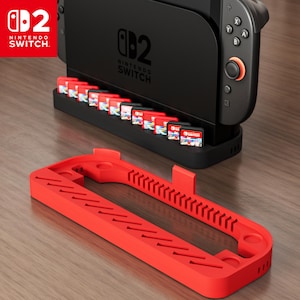 Może przedstawiać: Czarna konsola Nintendo Switch z uchwytem na kartridże do gier i czerwonym uchwytem na kartridże do gier. Logo Nintendo Switch jest widoczne na konsoli i kartridżach do gier. Czerwony uchwyt jest na pierwszym planie.
