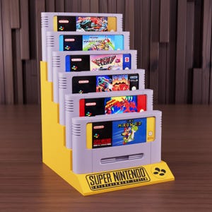 Puede incluir: Un expositor amarillo escalonado que contiene seis cartuchos de juegos de Super Nintendo. Los cartuchos son grises con etiquetas de juegos coloridas. El expositor tiene las palabras "SUPER NINTENDO ENTERTAINMENT SYSTEM" impresas.