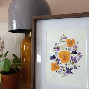 Op de afbeelding: Een ingelijst kunstwerk van geperste bloemen in tinten oranje, paars en blauw. De bloemen zijn gerangschikt op een witte achtergrond in een donkerbruine houten lijst. Een grijze lamp en een potplant staan op de achtergrond.