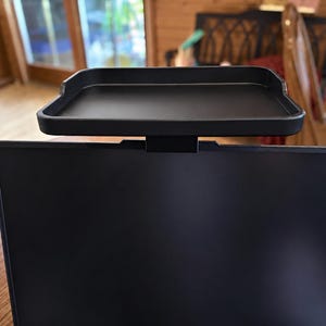 May include: A matte black monitor top shelf with a rectangular tray design and a mounting bracket attached to the centre, positioned securely on the top edge of a flat screen computer monitor with a dark, blank display screen.