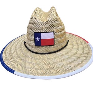 May include: A straw hat with a wide brim, featuring a Texas flag patch. The brim is trimmed with blue, white, and red stripes. A black cord encircles the hat's crown. This hat is suitable for outdoor wear.