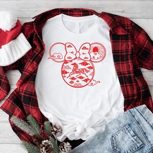 May include: White t-shirt with a red graphic design featuring a horse inside a Mickey Mouse-shaped design with clouds and a bow. A red and black plaid shirt, red and white beanie, and blue jeans are also in the image.