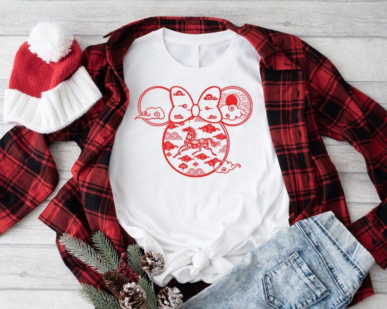 May include: White t-shirt with a red graphic design featuring a horse inside a Mickey Mouse-shaped design with clouds and a bow. A red and black plaid shirt, red and white beanie, and blue jeans are also in the image.