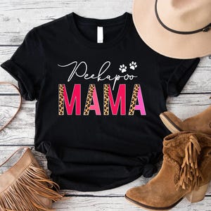 May include: Black t-shirt with the words "Peekapoo Mama" in white script and pink and leopard print block letters. The shirt is styled with a tan hat, brown boots, and a fringed purse.