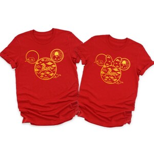 May include: Two red t-shirts with a gold design featuring a horse, clouds, and sun, arranged in a Mickey Mouse head shape. One shirt has a bow. The shirts are short-sleeved.