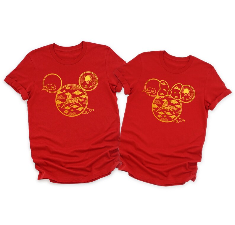 May include: Two red t-shirts with a gold design featuring a horse, clouds, and sun, arranged in a Mickey Mouse head shape. One shirt has a bow. The shirts are short-sleeved.