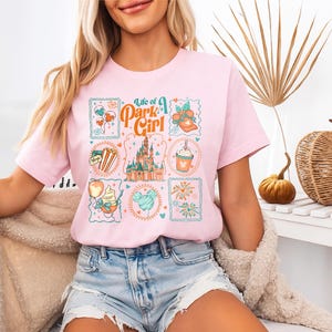 May include: A light pink t-shirt with the text "Life of a Park Girl" and various Disney-themed graphics. The graphics include a castle, balloons, and food items. The shirt is paired with denim shorts.