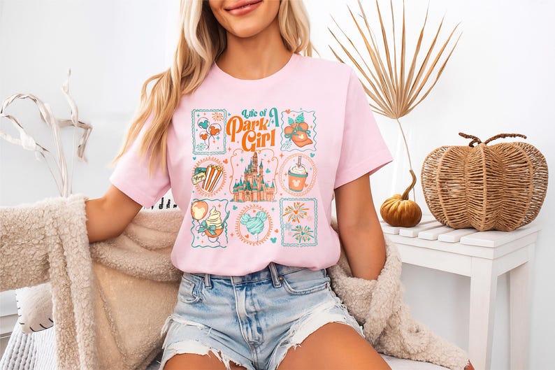 May include: A light pink t-shirt with the text "Life of a Park Girl" and various Disney-themed graphics. The graphics include a castle, balloons, and food items. The shirt is paired with denim shorts.