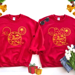 May include: Two red sweatshirts with gold designs. One features a Mickey Mouse head with a horse and cloud design, the other a Minnie Mouse head with a bow and similar design. Sunglasses and flowers are next to the sweatshirts.