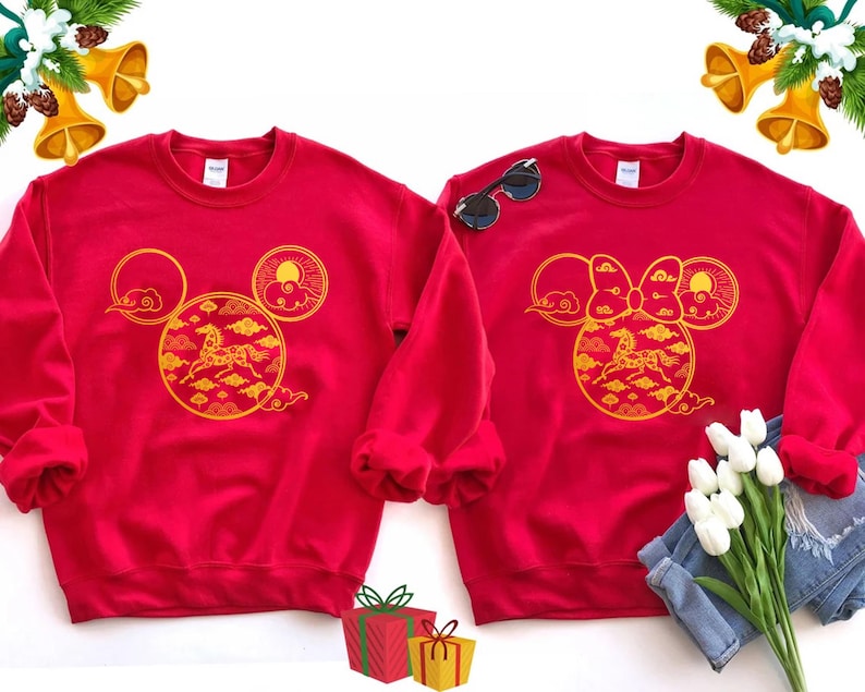 May include: Two red sweatshirts with gold designs. One features a Mickey Mouse head with a horse and cloud design, the other a Minnie Mouse head with a bow and similar design. Sunglasses and flowers are next to the sweatshirts.