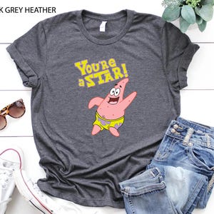 May include: Dark grey heather t-shirt featuring Patrick Star from Spongebob Squarepants. The shirt has the text "You're a STAR!" in yellow, with Patrick in his yellow shorts. Sunglasses, white sneakers, and blue jeans are also in the image.