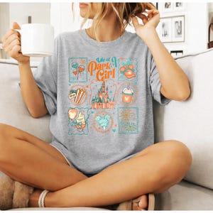 May include: A gray t-shirt with the text "Life of a Park Girl" and various colorful illustrations. The illustrations include a castle, balloons, popcorn, and a drink. The shirt is worn by a person sitting on a couch.