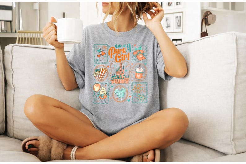 May include: A gray t-shirt with the text "Life of a Park Girl" and various colorful illustrations. The illustrations include a castle, balloons, popcorn, and a drink. The shirt is worn by a person sitting on a couch.