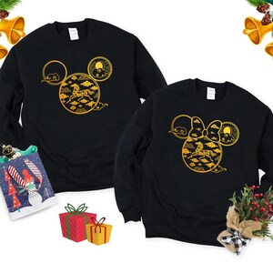 May include: Two black sweatshirts with a gold design featuring a horse, clouds, and sun, arranged in a Mickey Mouse head shape. Festive decorations, including bells and gifts, surround the sweatshirts.