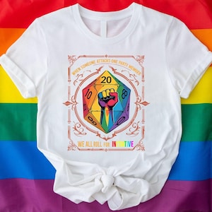May include: White t-shirt featuring a colorful D20 die with a rainbow fist graphic. Text reads "WHEN SOMEONE ATTACKS ONE PARTY MEMBER" and "WE ALL ROLL FOR INITIATIVE". The shirt is displayed against a rainbow flag.