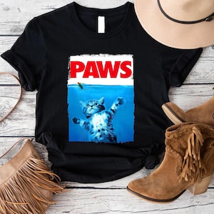 May include: Black t-shirt featuring a parody of the movie "Jaws" with a cat in the water and the word "PAWS" in red. The cat is blue and white, with its paws outstretched. The shirt is displayed on a wooden surface.