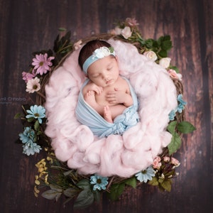 Puede incluir: Una recién nacida duerme en un nido hecho de ramitas y material rosa y esponjoso. El nido está decorado con flores artificiales y vegetación. La bebé lleva un arrullo azul claro y una diadema blanca con una flor.