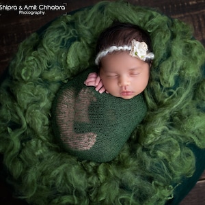 May include: A newborn baby is swaddled in a green knitted wrap and sleeping peacefully in a green fluffy nest. The baby is wearing a white headband with a small white flower.