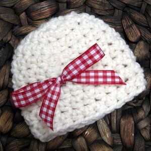 May include: A white crocheted baby hat with a red and white gingham ribbon tied in a bow.
