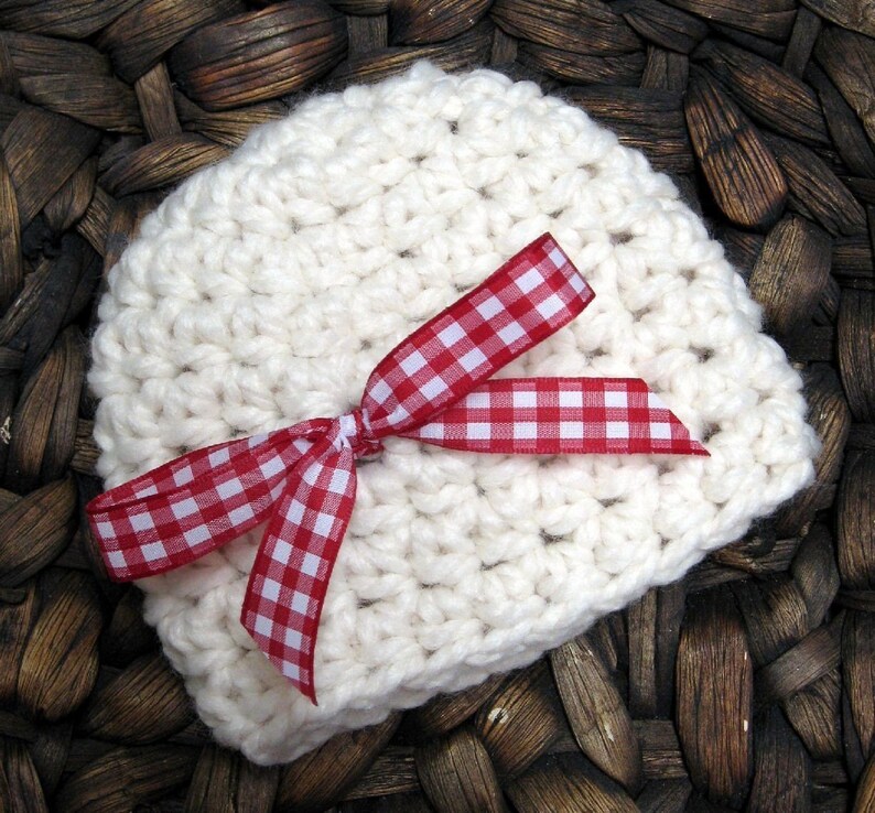 May include: A white crocheted baby hat with a red and white gingham ribbon tied in a bow.