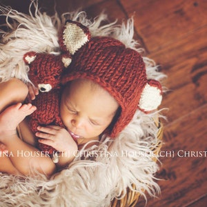 May include: A newborn baby wearing a knitted fox hat with white ears and a brown knitted fox toy. The baby is sleeping in a basket lined with white fur.