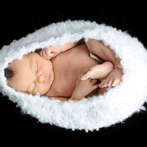 May include: A newborn baby sleeping peacefully in a white crocheted nest. The baby is curled up with their arms and legs tucked in, and their face is serene.
