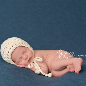 May include: A newborn baby wearing a white crocheted bonnet sleeps on a blue blanket. The baby is curled up with their arms and legs tucked in. The baby's face is visible and they have their eyes closed.