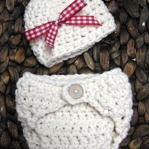 May include: A white crocheted baby hat with a red and white gingham bow. The hat is lying on a woven brown basket.