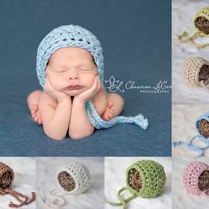 May include: A baby wearing a light blue crocheted bonnet with ties, resting on blue fabric. Other bonnets in green, beige, brown, white, and pink are shown. The image is a product shot for baby photography props. The bonnets are likely made of wool or cotton.
