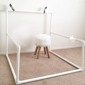 May include: White PVC pipe backdrop stand with a white furry stool in the center. The stand is assembled in a square shape with a top bar for hanging backdrops.