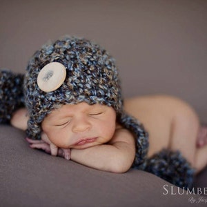 Op de afbeelding: Een slapende baby met een blauwe, bruine en witte gehaakte muts met oorflappen en een grote knoop. De baby ligt op een grijze ondergrond. De tekst "SLUMBERLINGS" is rechtsonder zichtbaar.