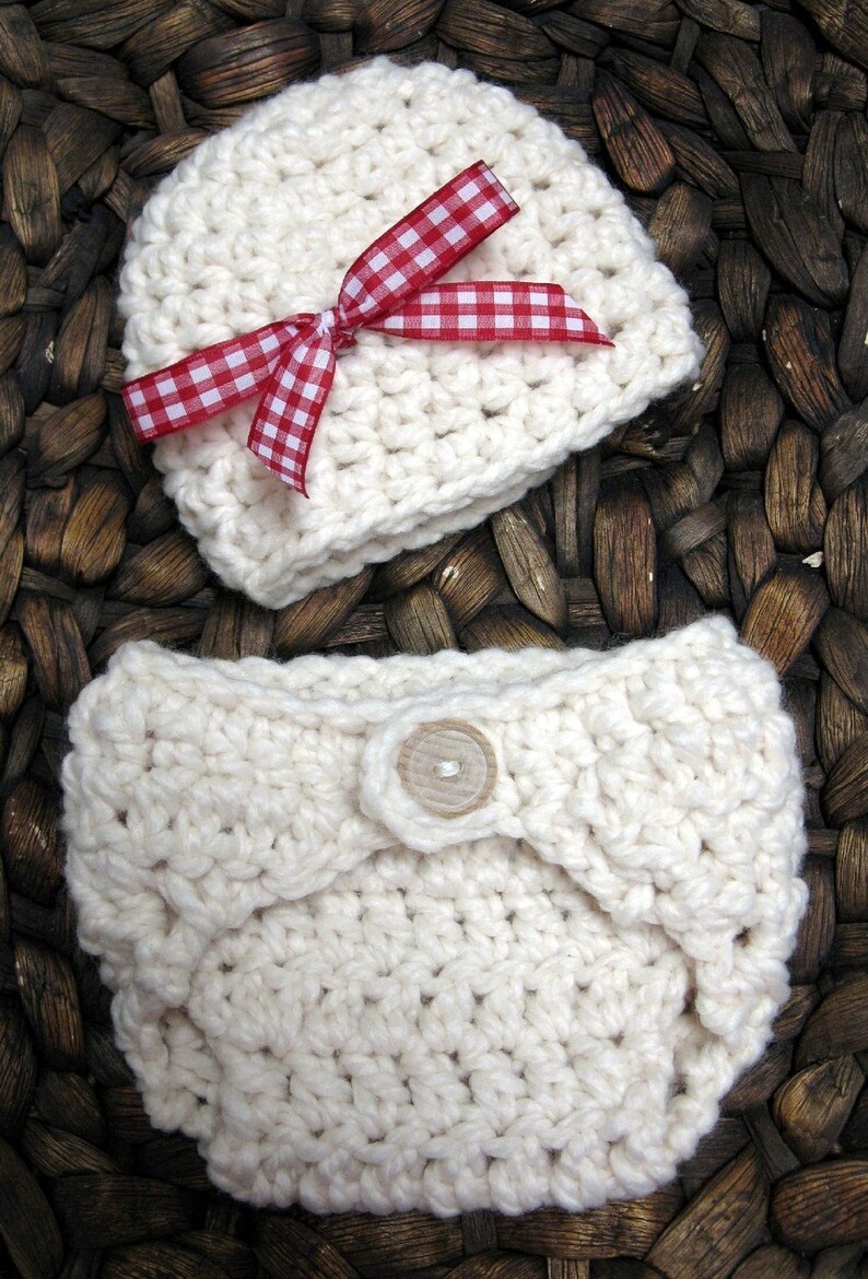 May include: A white crocheted baby hat with a red and white gingham bow and a white crocheted diaper with a wooden button.