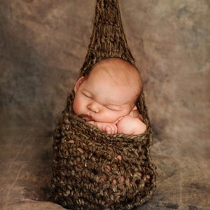 May include: A newborn baby sleeping soundly in a brown crocheted hammock. The baby is swaddled in the hammock and has its arms tucked in.