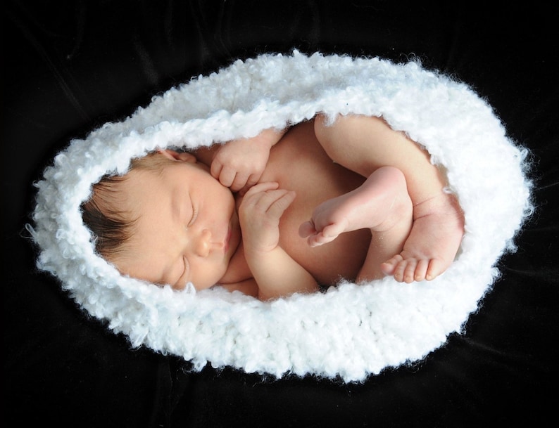 May include: A newborn baby sleeping peacefully in a white crocheted nest on a black background.
