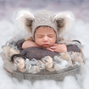 Puede incluir: Un bebé recién nacido con un gorro gris de punto con orejas peludas, durmiendo en una cesta esponjosa gris y blanca.