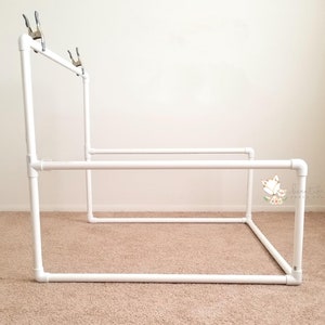 May include: White PVC pipe backdrop stand with clamps for photography. The stand is assembled in a rectangular shape.
