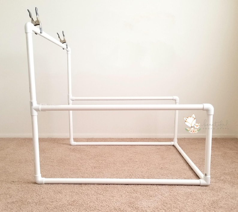 May include: White PVC pipe backdrop stand with clamps for photography. The stand is assembled in a rectangular shape.
