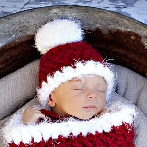 May include: A red and white crocheted Santa hat with a white pom-pom on top. The hat is worn by a sleeping baby.