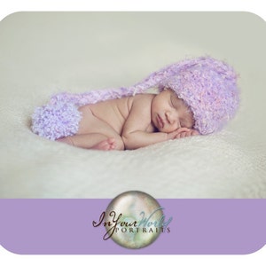 May include: A newborn baby sleeping on a white blanket, wearing a soft, lavender knitted hat with a fluffy pom-pom. The baby is curled up in a peaceful slumber.