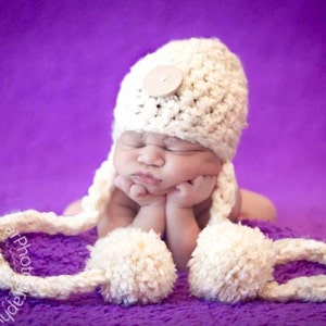 May include: A white crocheted hat with a pom-pom on the end of the long tail. The hat has a button on the top and is worn by a baby sleeping on a purple background.