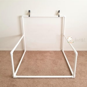 May include: White PVC pipe backdrop stand for photography. The stand is assembled in a square shape with a top bar and two cross bars. The stand is secured to the wall with clamps.