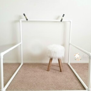 May include: White PVC pipe backdrop stand with a white faux fur stool in the center. The stand is assembled in a rectangular shape with two horizontal bars and two vertical bars.